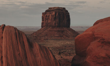 Formaciones rocosas de arenisca roja en el desierto, con un butte columnar al fondo al atardecer