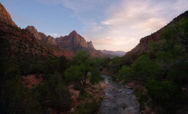 Cañón con río entre acantilados de roca roja, vegetación verde y montañas al fondo al atardecer