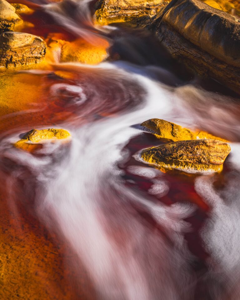 Long exposure photograph of flowing water over mineral rocks with red, orange and white tones.