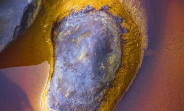 Fotografía abstracta de una formación mineral con núcleo claro y bordes oxidados en tonos amarillos, ocres y azulados.