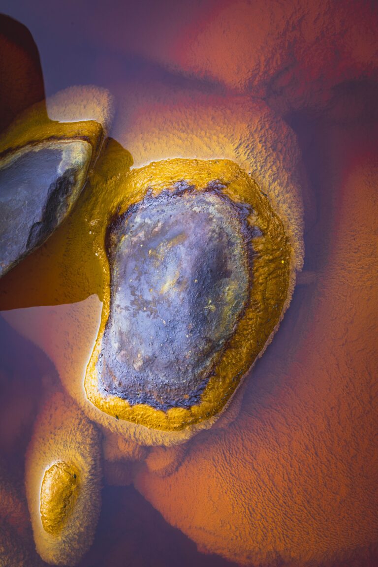 Fotografía abstracta de una formación mineral con núcleo claro y bordes oxidados en tonos amarillos, ocres y azulados.