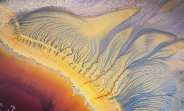 Fotografía aérea abstracta de sedimentos minerales con patrones orgánicos en tonos amarillos, azules y anaranjados.