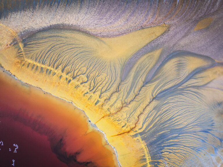 Fotografía aérea abstracta de sedimentos minerales con patrones orgánicos en tonos amarillos, azules y anaranjados.
