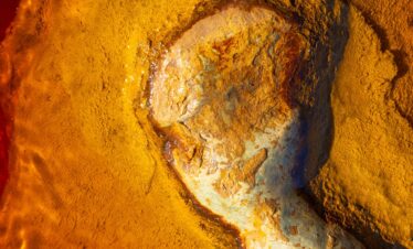 Abstract aerial photograph of mineral-rich terrain with golden, ochre and blue textures forming organic patterns.