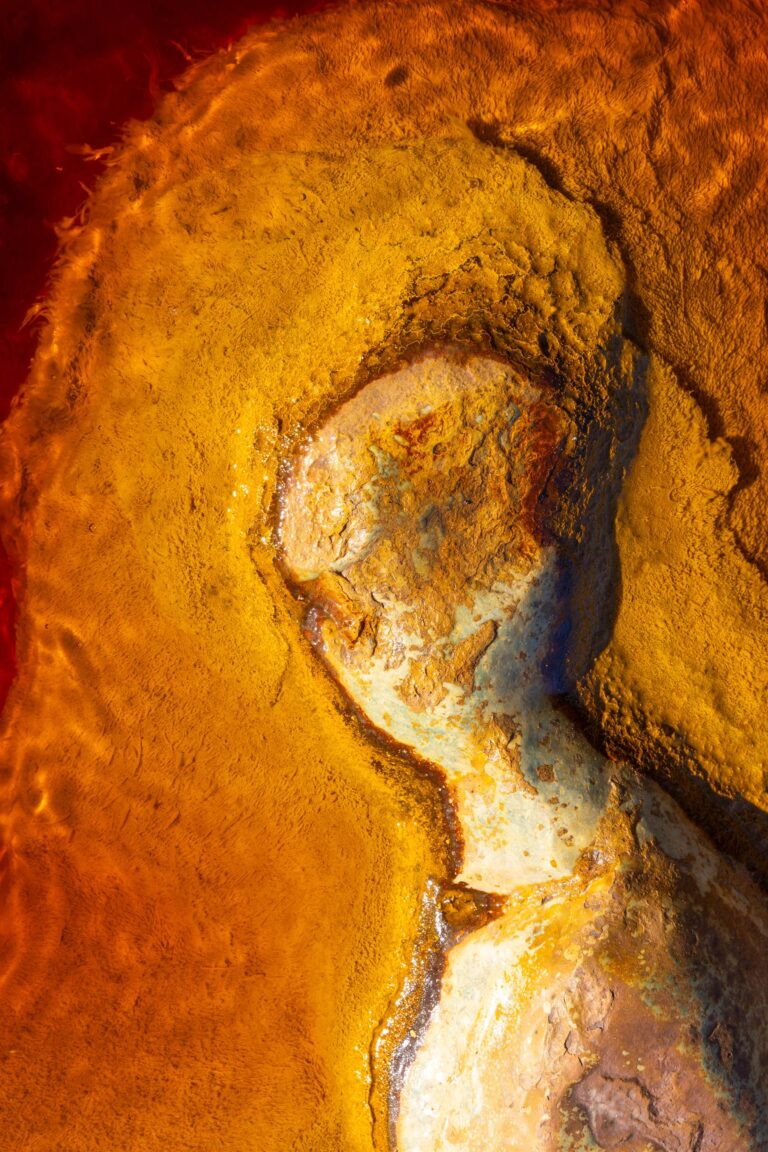 Abstract aerial photograph of mineral-rich terrain with golden, ochre and blue textures forming organic patterns.