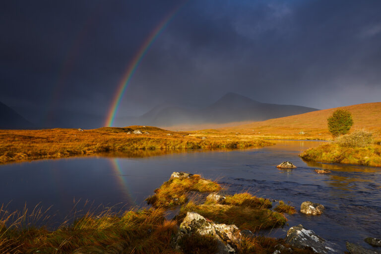 Arcoíris sobre un valle escocés con río, montaña al fondo y luz dorada iluminando la pradera