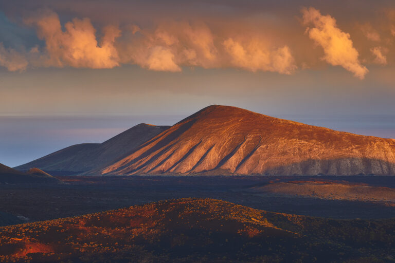 Volcanes de Lanzarote al atardecer con luz dorada iluminando sus laderas y nubes teñidas de naranja en el cielo