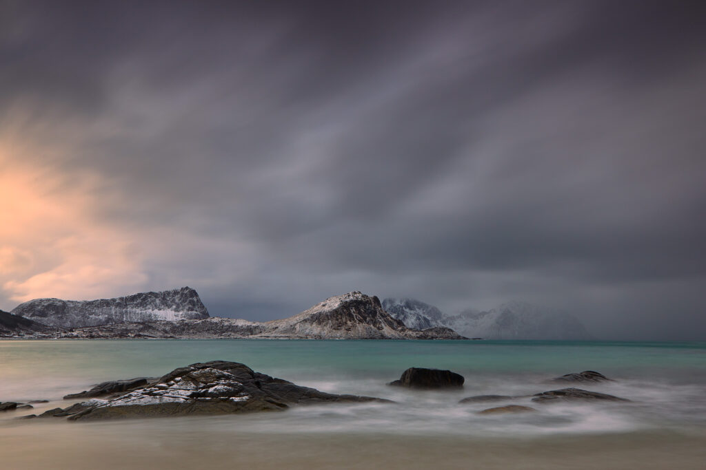 Playa ártica con rocas en primer plano, aguas turquesas y montañas nevadas al fondo bajo cielo nublado