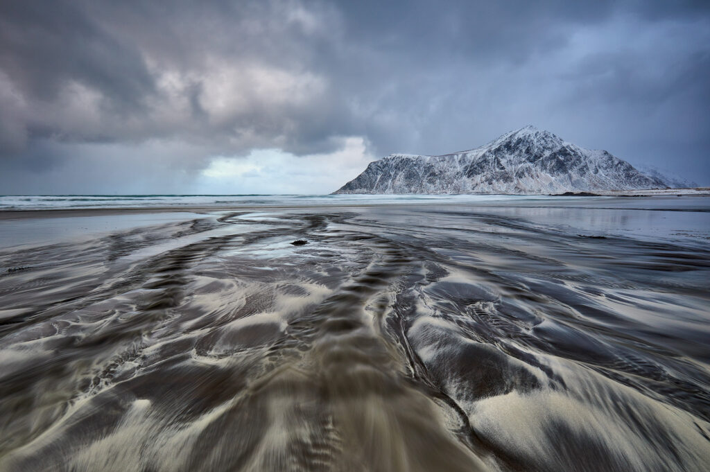 Playa con arena ondulante y montaña nevada al fondo bajo cielo nublado