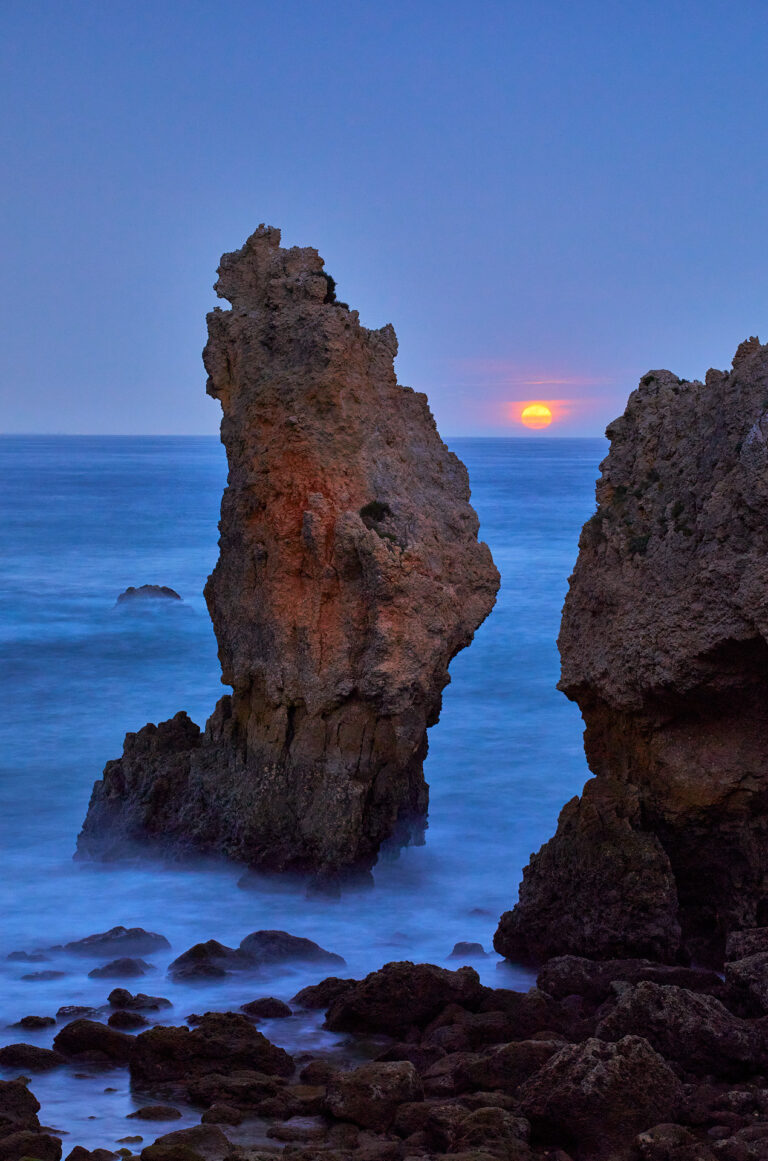 Formación rocosa en el mar al atardecer con sol naranja en horizonte y aguas azules
