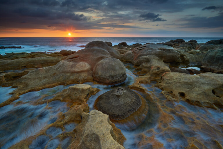 Rocas graníticas en la costa al atardecer, con olas azules fluyendo entre formaciones geológicas durante la puesta de sol