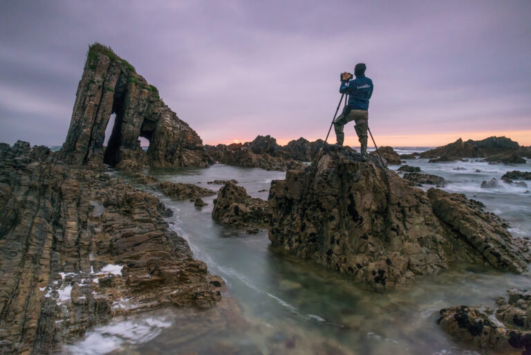 Fotógrafo sobre roca en costa rocosa al atardecer, acantilados hexagonales y mar con olas de fondo