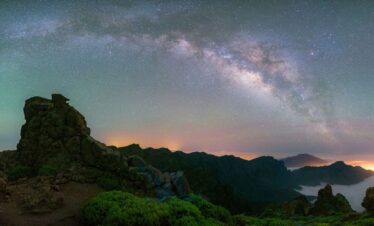 Vía Láctea sobre panorámica nocturna de La Palma con andenes de piedra, mar de nubes y volcanes iluminados al atardecer