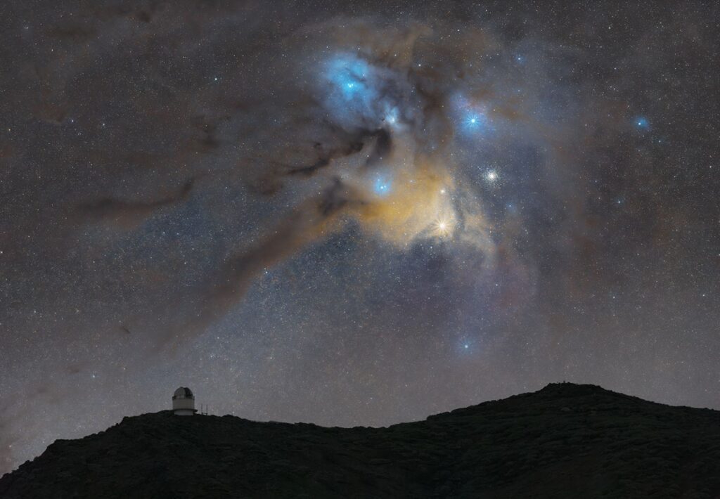 Cielo nocturno estrellado sobre silueta de montaña con observatorio astronómico, nebulosa Rho Ofiuchi azul y amarilla