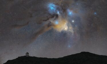 Cielo nocturno estrellado sobre silueta de montaña con observatorio astronómico, nebulosa Rho Ofiuchi azul y amarilla