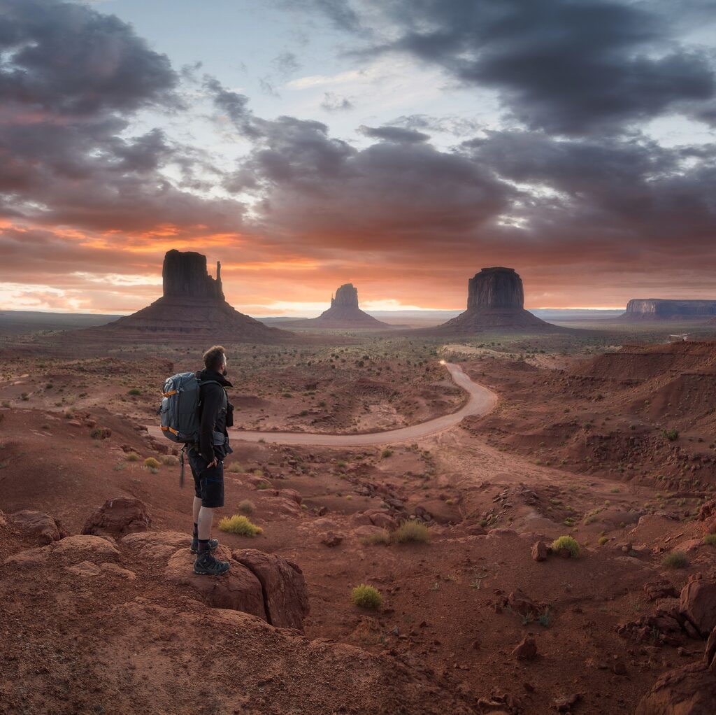 Senderista con mochila observa los monumentos de Monument Valley al atardecer, con camino serpenteante en el desierto