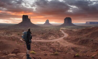 Senderista con mochila observa los monumentos de Monument Valley al atardecer, con camino serpenteante en el desierto