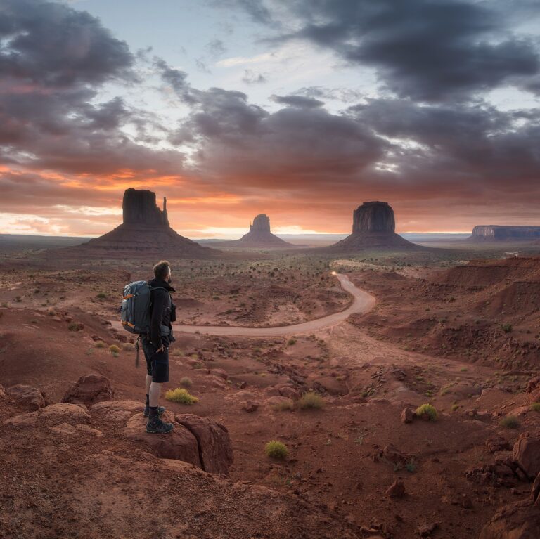Senderista con mochila observa los monumentos de Monument Valley al atardecer, con camino serpenteante en el desierto