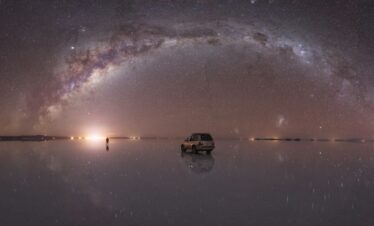 Vía Láctea arqueada sobre el Salar de Uyuni con reflejo perfecto en agua, vehículo y personas bajo cielo estrellado