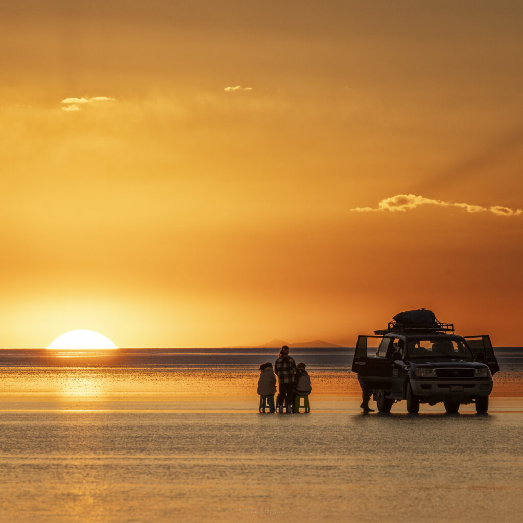 Grupo de personas junto a un vehículo 4x4 observan la puesta de sol en el Salar de Uyuni, con el cielo teñido de naranja y oro