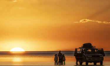 Grupo de personas junto a un vehículo 4x4 observan la puesta de sol en el Salar de Uyuni, con el cielo teñido de naranja y oro