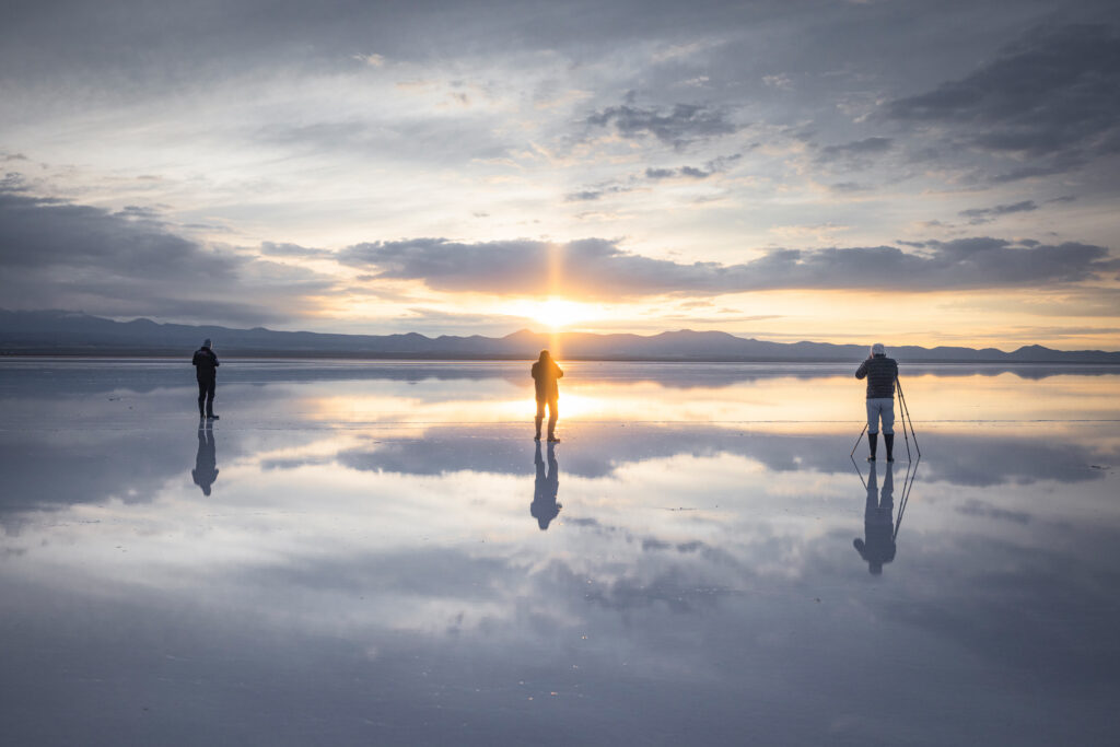 Tres personas en bicicleta sobre el espejo de agua del Salar de Uyuni al atardecer, con montañas al fondo y reflejo perfecto del cielo.