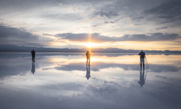 Tres personas en bicicleta sobre el espejo de agua del Salar de Uyuni al atardecer, con montañas al fondo y reflejo perfecto del cielo.