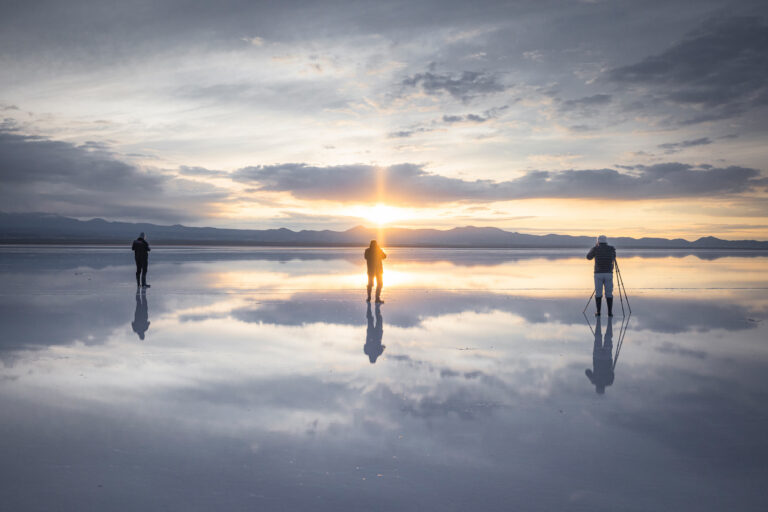 Tres personas en bicicleta sobre el espejo de agua del Salar de Uyuni al atardecer, con montañas al fondo y reflejo perfecto del cielo.