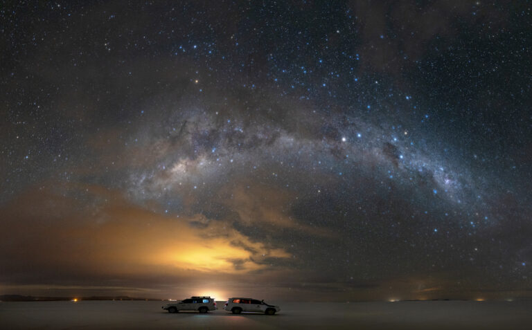 Dos vehículos en el Salar de Uyuni bajo la Vía Láctea y resplandor crepuscular en el horizonte