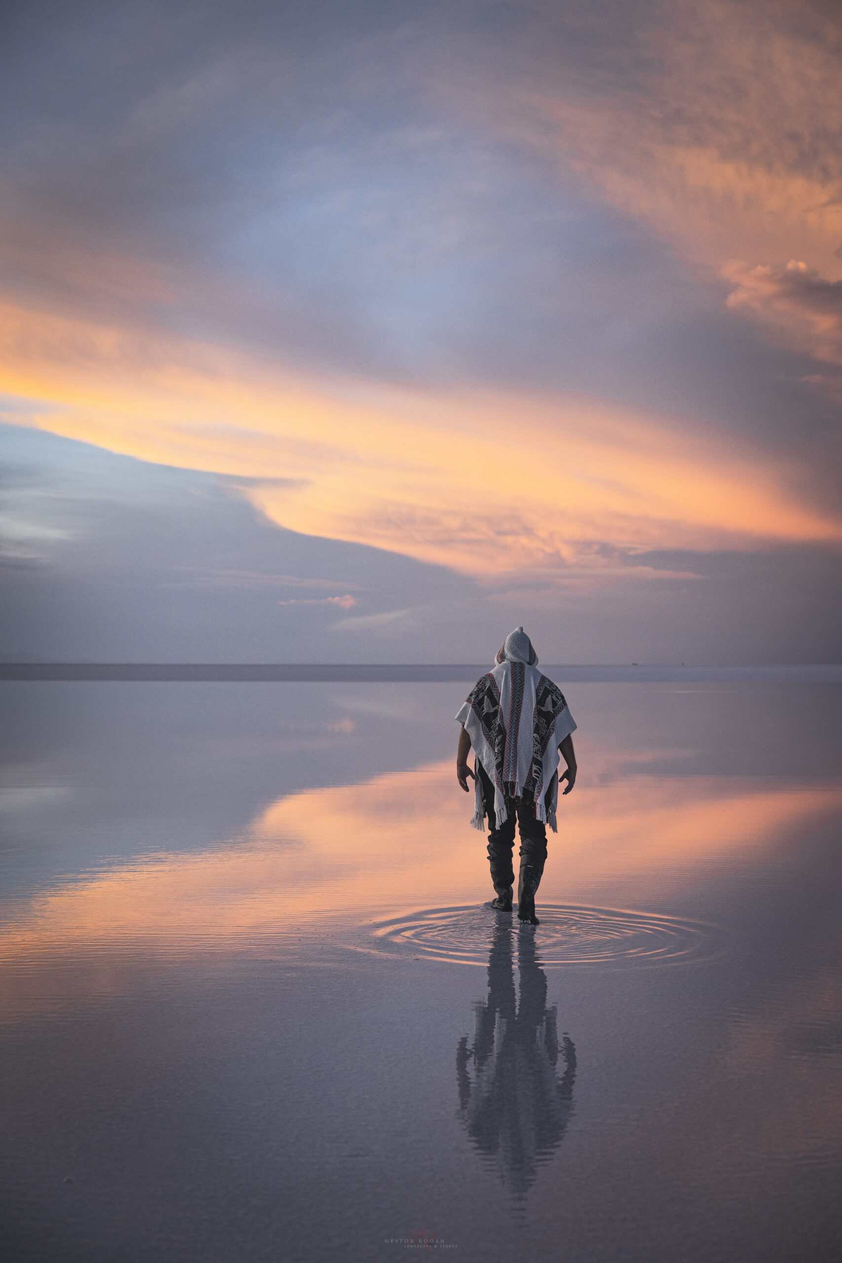 Persona en poncho de pie en el Salar de Uyuni al atardecer, reflejada en agua espejo con ondas concéntricas