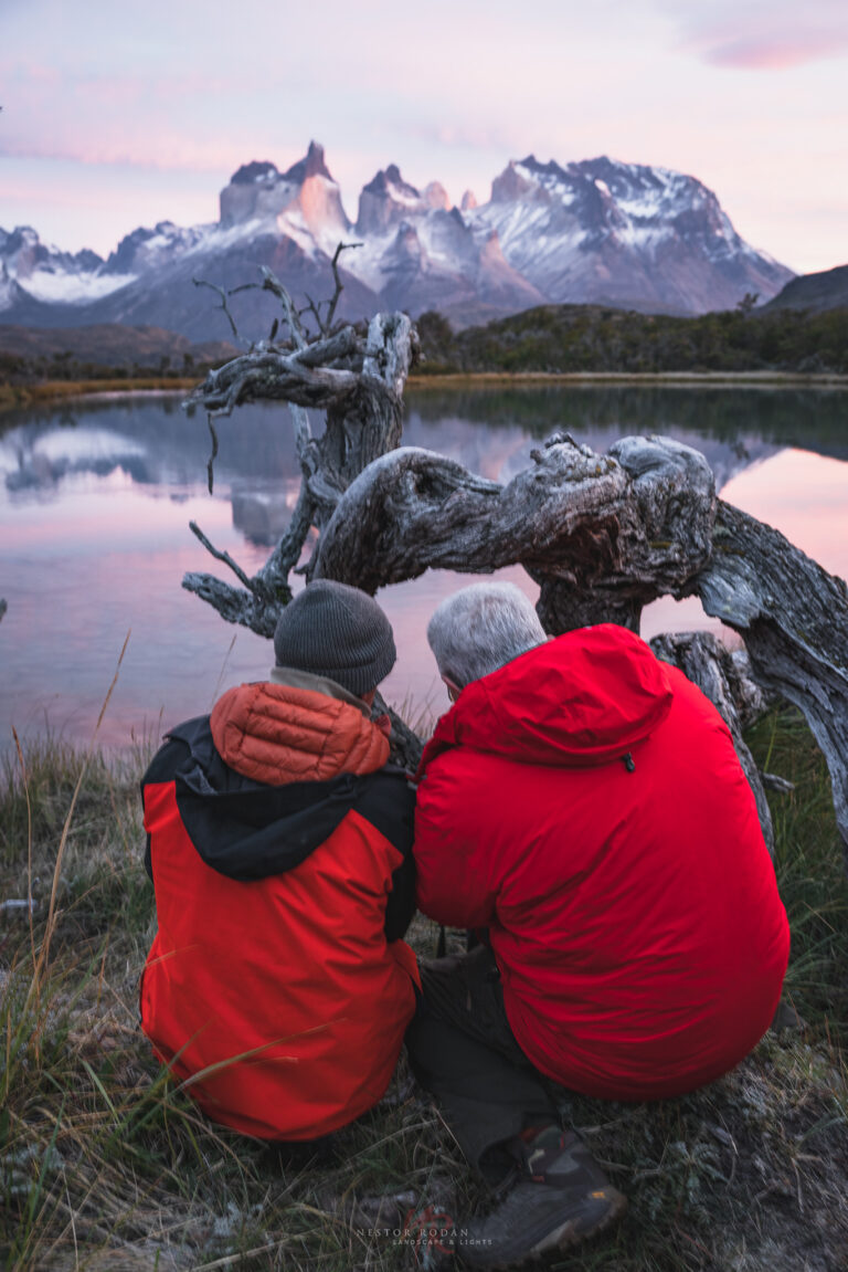 Dos viajeros de espaldas en chaquetas rojas observan las montañas de Torres del Paine reflejadas en un lago de la Patagonia