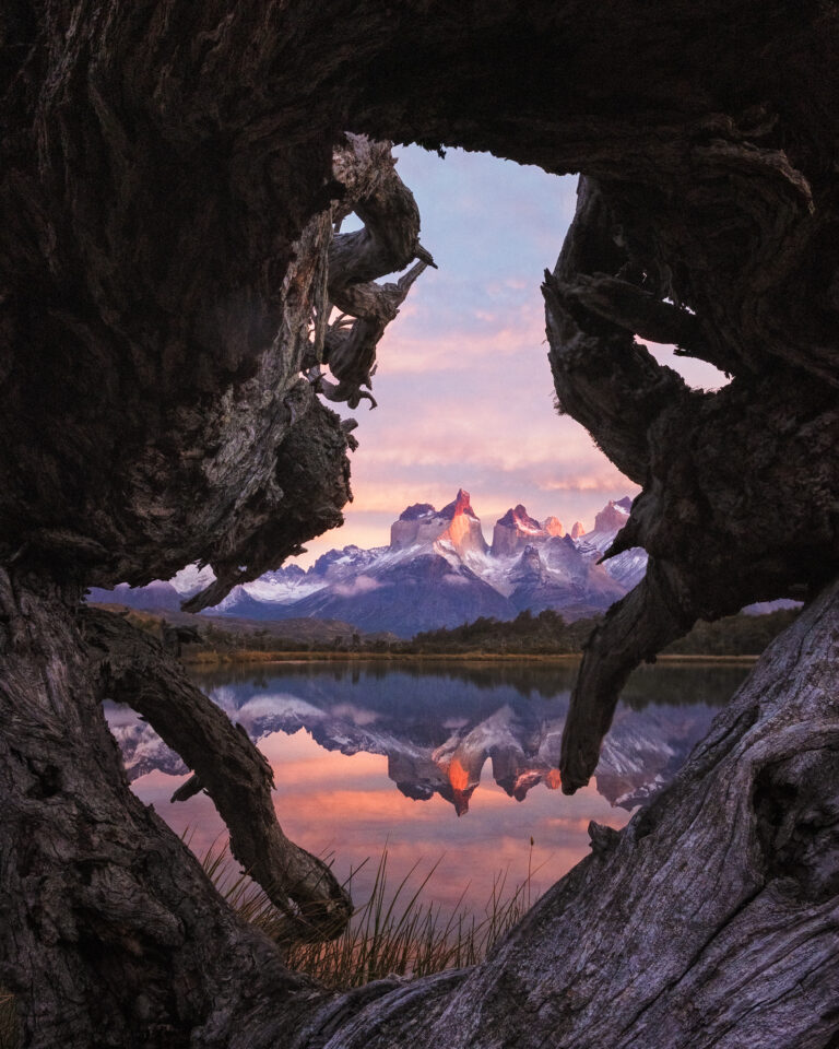 Vista desde cueva de roca erosionada hacia picos montañosos nevados reflejados en lago al atardecer en Patagonia