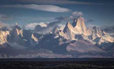 Picos nevados de granito en la Patagonia argentina al atardecer, con meseta desértica en primer plano y cielo nublado.