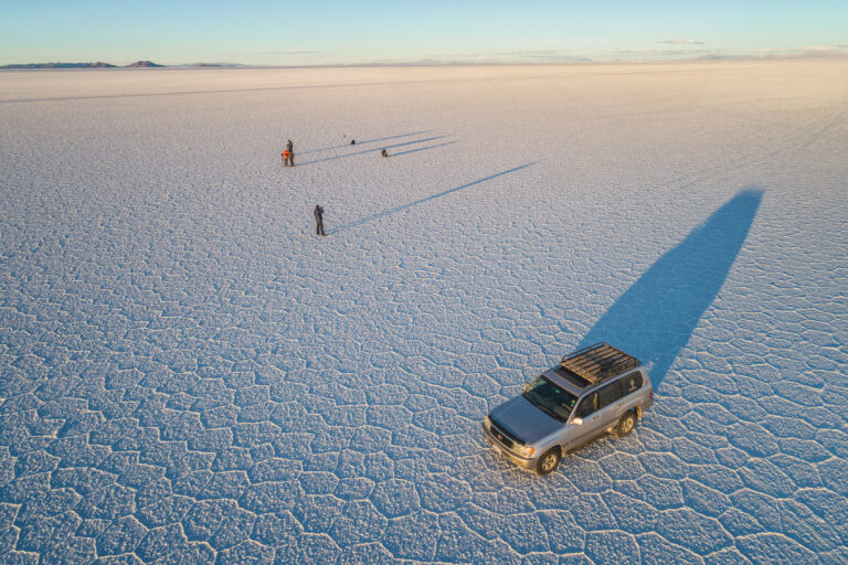 Vehículo 4x4 con sombra alargada en el Salar de Uyuni, con personas caminando en la planicie salina de Bolivia