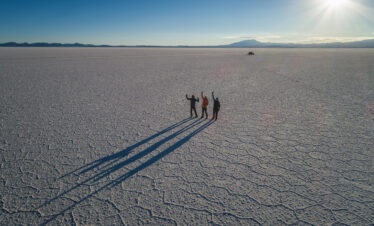 Tres expedicionarios en el vasto Salar de Uyuni, Bolivia, con largas sombras proyectadas sobre la superficie blanca del salares y montañas al horizonte.