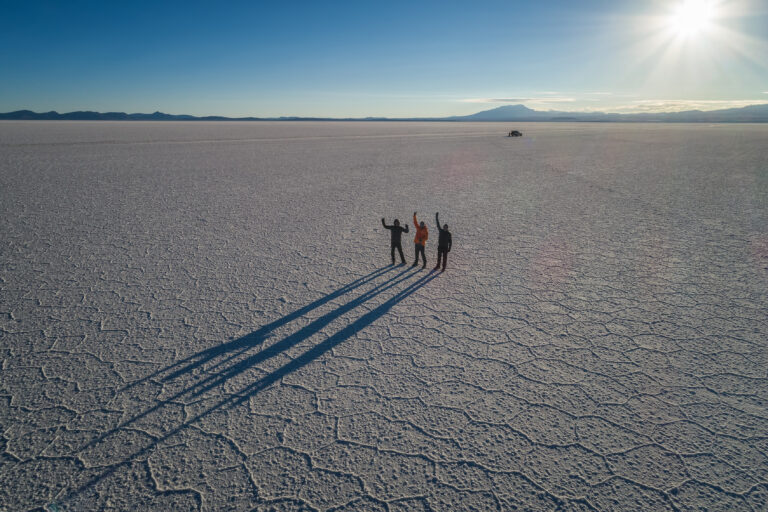 Tres expedicionarios en el vasto Salar de Uyuni, Bolivia, con largas sombras proyectadas sobre la superficie blanca del salares y montañas al horizonte.