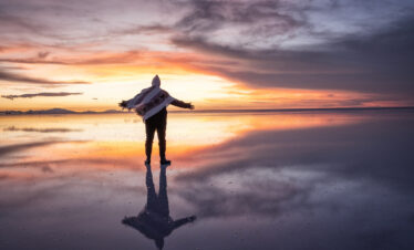Surfista con tabla en el Salar de Uyuni al atardecer, reflejado en el espejo de agua salada durante la estación húmeda.