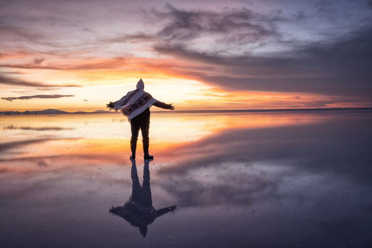 Surfista con tabla en el Salar de Uyuni al atardecer, reflejado en el espejo de agua salada durante la estación húmeda.