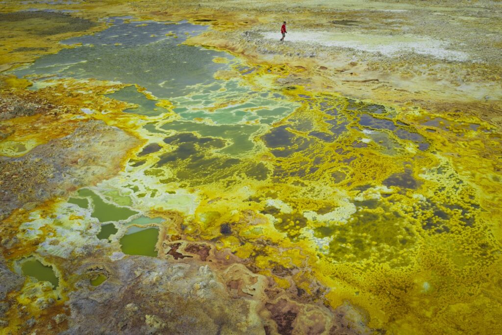 Termas geotérmicas con aguas minerales amarillas y verdes en Etiopía, con un explorador caminando entre fumarolas
