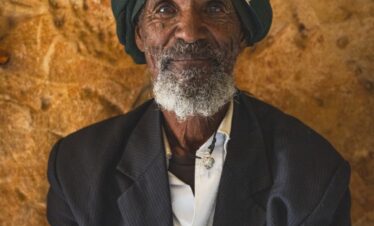 Retrato de anciano etíope con turbante azul y traje oscuro contra pared de tierra ocre