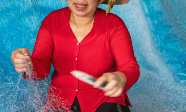 Mujer vietnamita con áo dài rojo y sombrero cónico (nón lá) reparando red de pesca sobre fondo azul