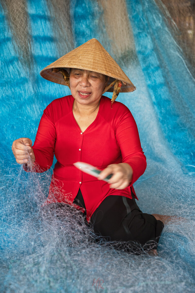 Mujer vietnamita con áo dài rojo y sombrero cónico (nón lá) reparando red de pesca sobre fondo azul