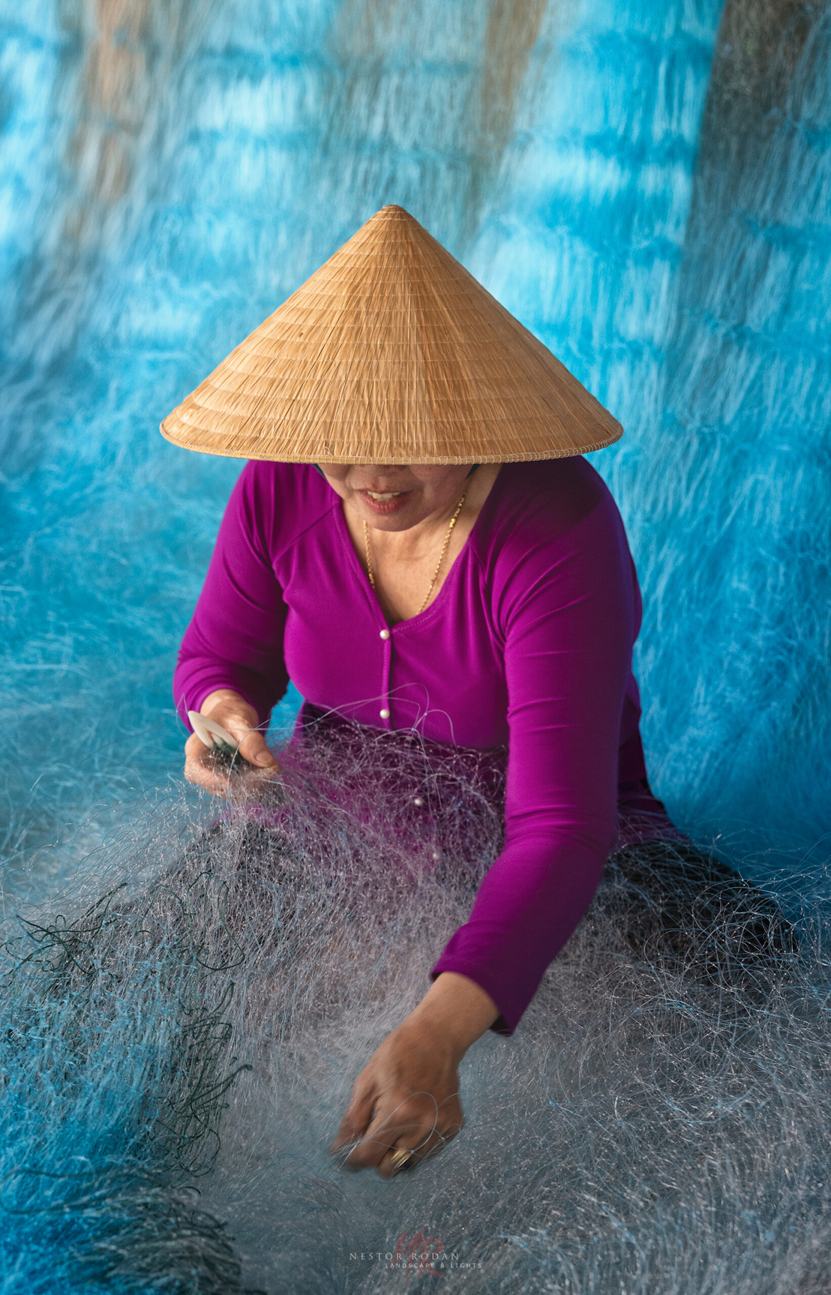 Mujer vietnamita con áo dài magenta tejiendo redes de pesca en el Delta del Mekong, frente a agua azul turquesa