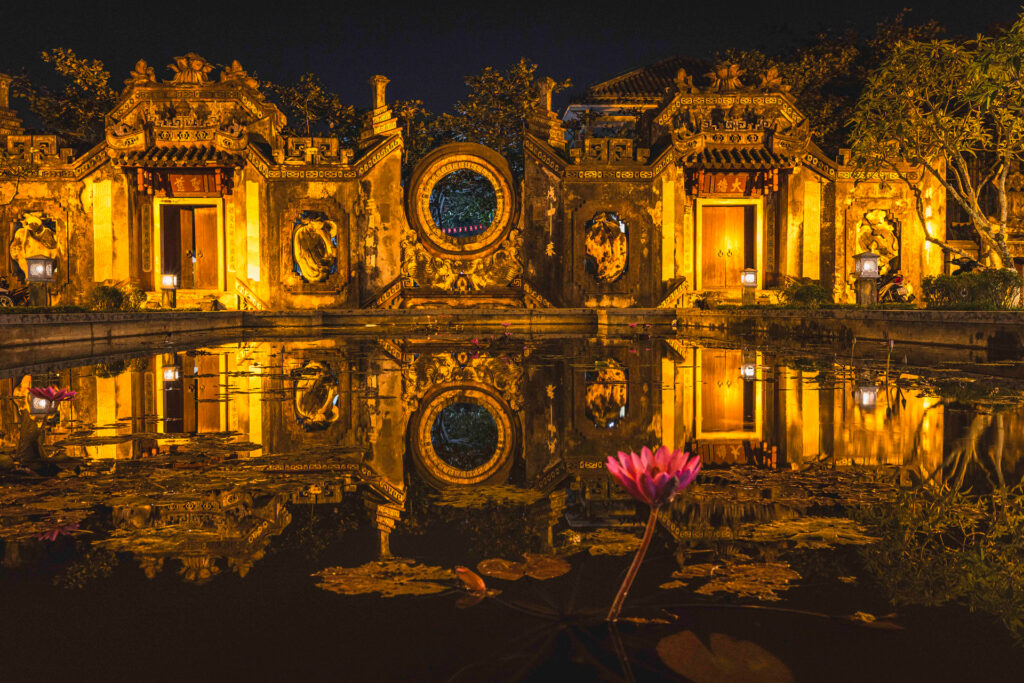 Templo asiático iluminado al atardecer con reflejo en agua y flor de loto rosa en primer plano