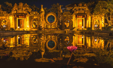 Templo asiático iluminado al atardecer con reflejo en agua y flor de loto rosa en primer plano