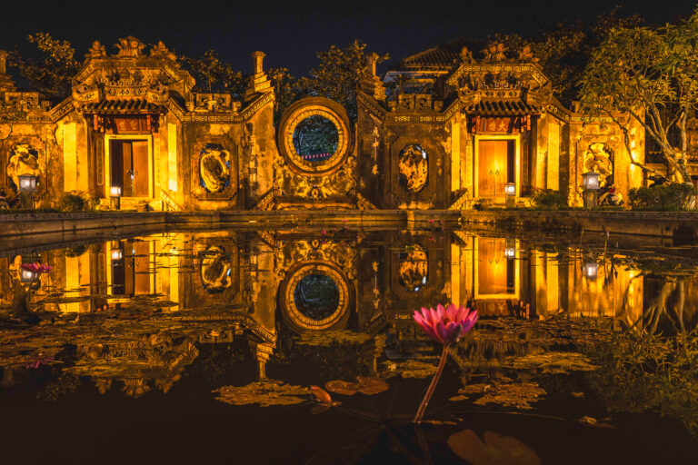 Templo asiático iluminado al atardecer con reflejo en agua y flor de loto rosa en primer plano