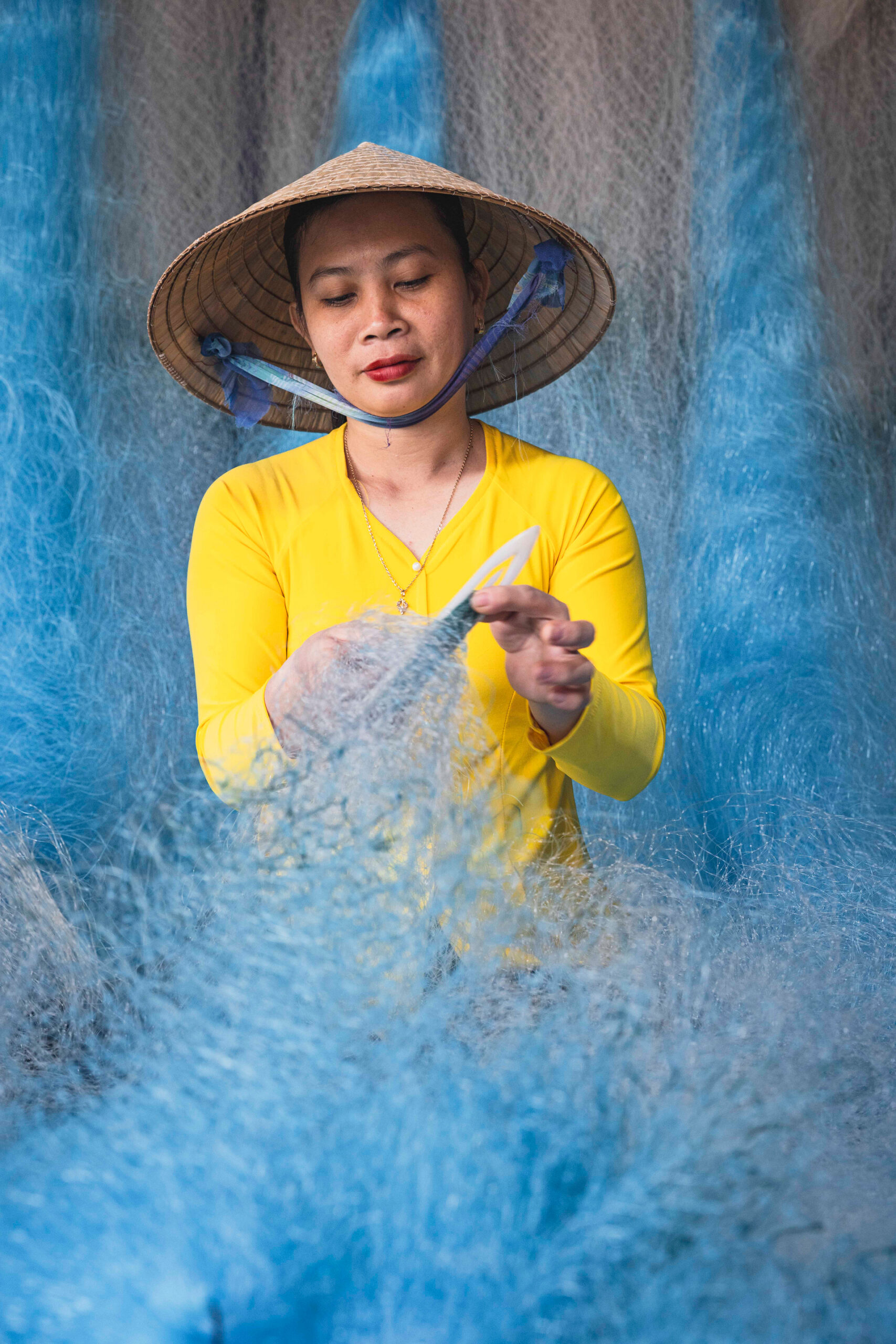 Mujer vietnamita en áo dài amarillo con sombrero cónico tejiendo una red de pesca azul