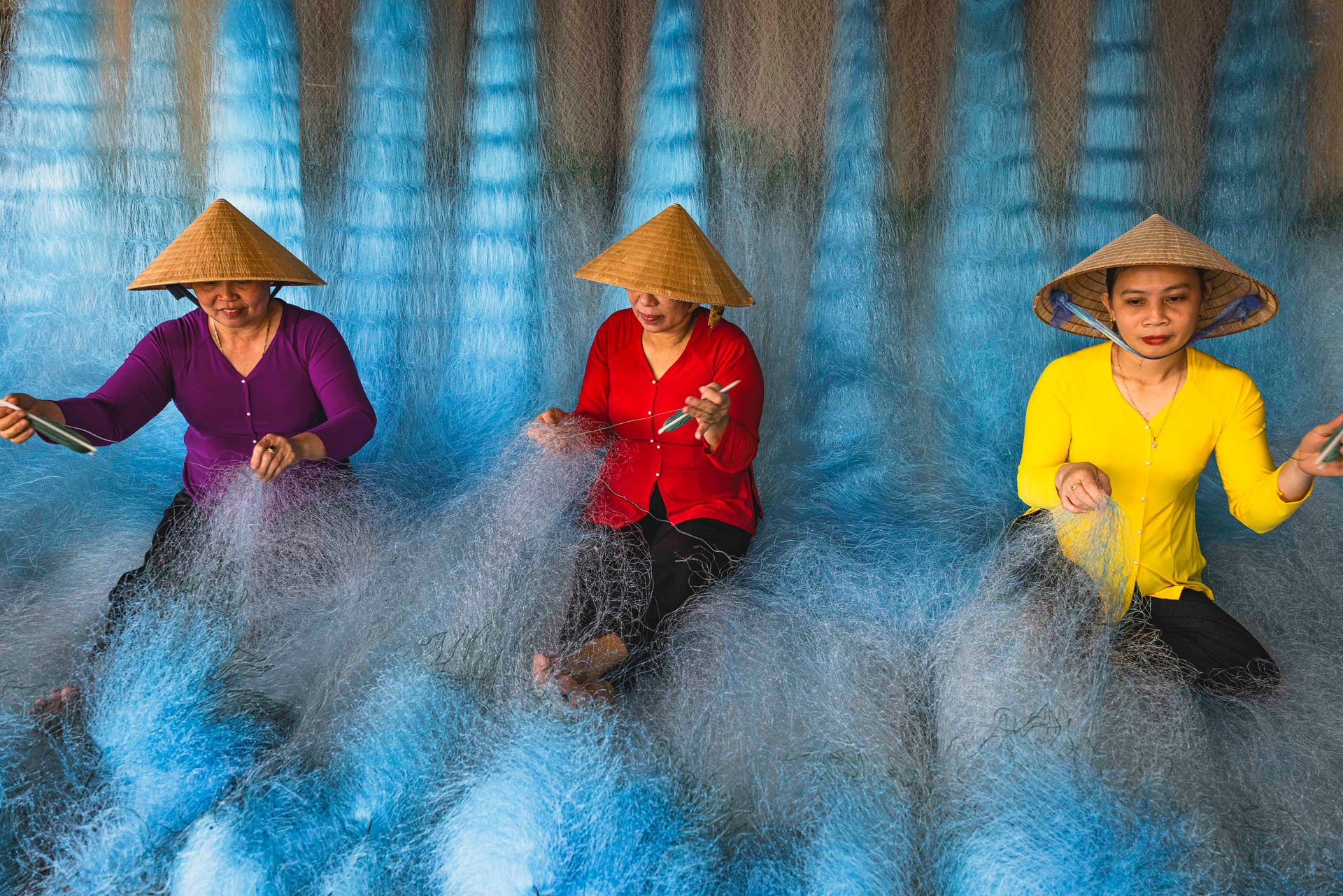 Tres mujeres vietnamitas con áo dài y sombreros cónicos tejen redes de pesca en el Delta del Mekong