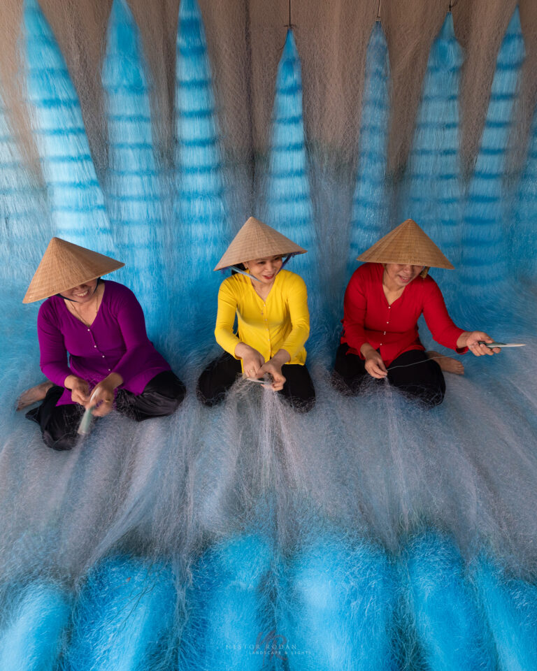 Tres mujeres vietnamitas con áo dài de colores tejen redes de pesca sentadas frente a un mural azul