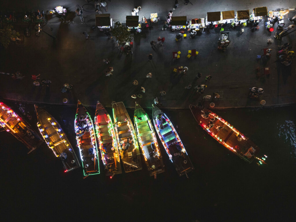Barcas de madera iluminadas con luces de colores amarradas en el río durante la noche, con mercado y gente en el muelle